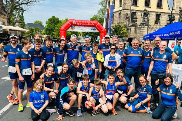 Eine große Gruppe von jungen und mittelalten Athlet*innen in blauen Funktionsshirts von DOWE. Sie posieren sitzend und stehend vor einer Zielmarkierung, vermutlich bei einem Stadtlauf