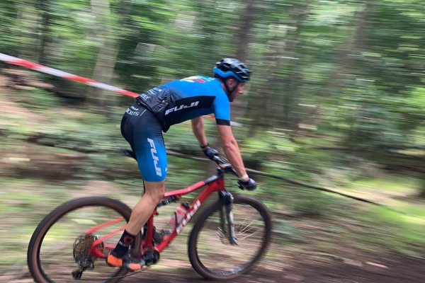 Ein Mountainbike-Fahrer steht auf seinem roten Fully-Bike und rast einen bewaldeten Hang hinab. Er trägt ein blaues Trikot mit der Aufschift "FUJI Rockets" von DOWE.