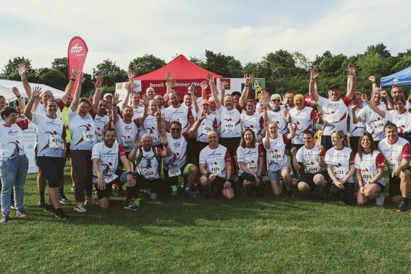 Ein Sportteam "VBK Verkehrsbetriebe Karlsruhe" mit weißen Running-Shirts von DOWE Sportswear posiert mit erhobenen Armen gut gelaunt vor Zelten im Freien.