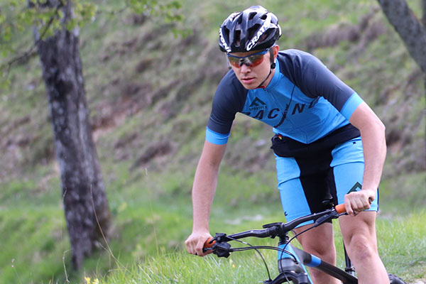 Ein Mountainbiker radelt im stehen. Er trägt blaues Trikot und blaue Short von DOWE Sportswear, die beide mit einem Custom-Logo des Vereins Sportfex bedruckt wurden. Im Hintergrund ist eine Wiese zu erkenne.