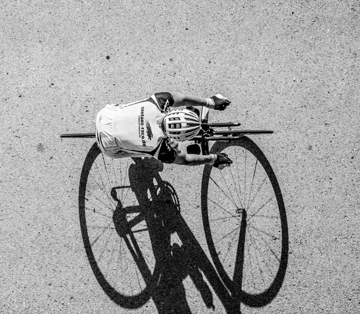 Ein Rennradfahrer in der Mittagssonnen aus der Vogelperspektive zu sehen. Das Bild ist eine schwarz-weiß-Aufnahme und der Radler wirft einen starken Schatten, welcher nach unten hin entweicht.