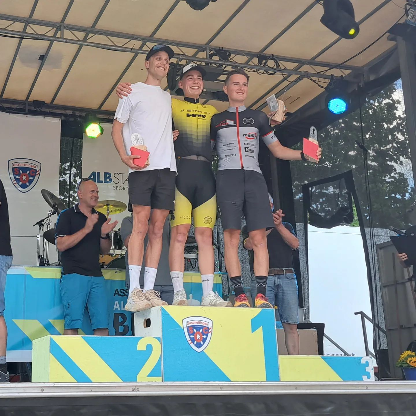 Drei Radsport-Athleten auf einer Tribüne. Sie halten Trophäen oder Geschenke. Der mittlere trägt ein gelbes Trikot der Firma DOWE Sportswear und und umarmt seine Nachbarn. Alle drei lächeln in die Kamera, welche von unten "hinaufblickt".