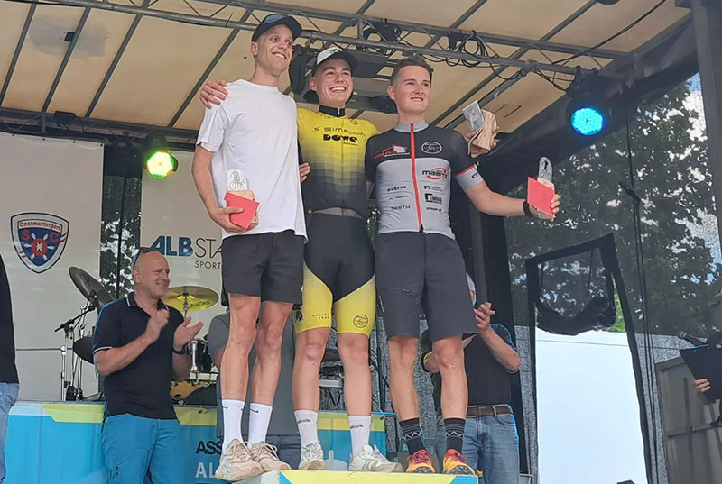 Drei Radsport-Athleten auf einer Tribüne. Sie halten Trophäen oder Geschenke. Der mittlere trägt ein gelbes Trikot der Firma DOWE Sportswear und und umarmt seine Nachbarn. Alle drei lächeln in die Kamera, welche von unten "hinaufblickt".