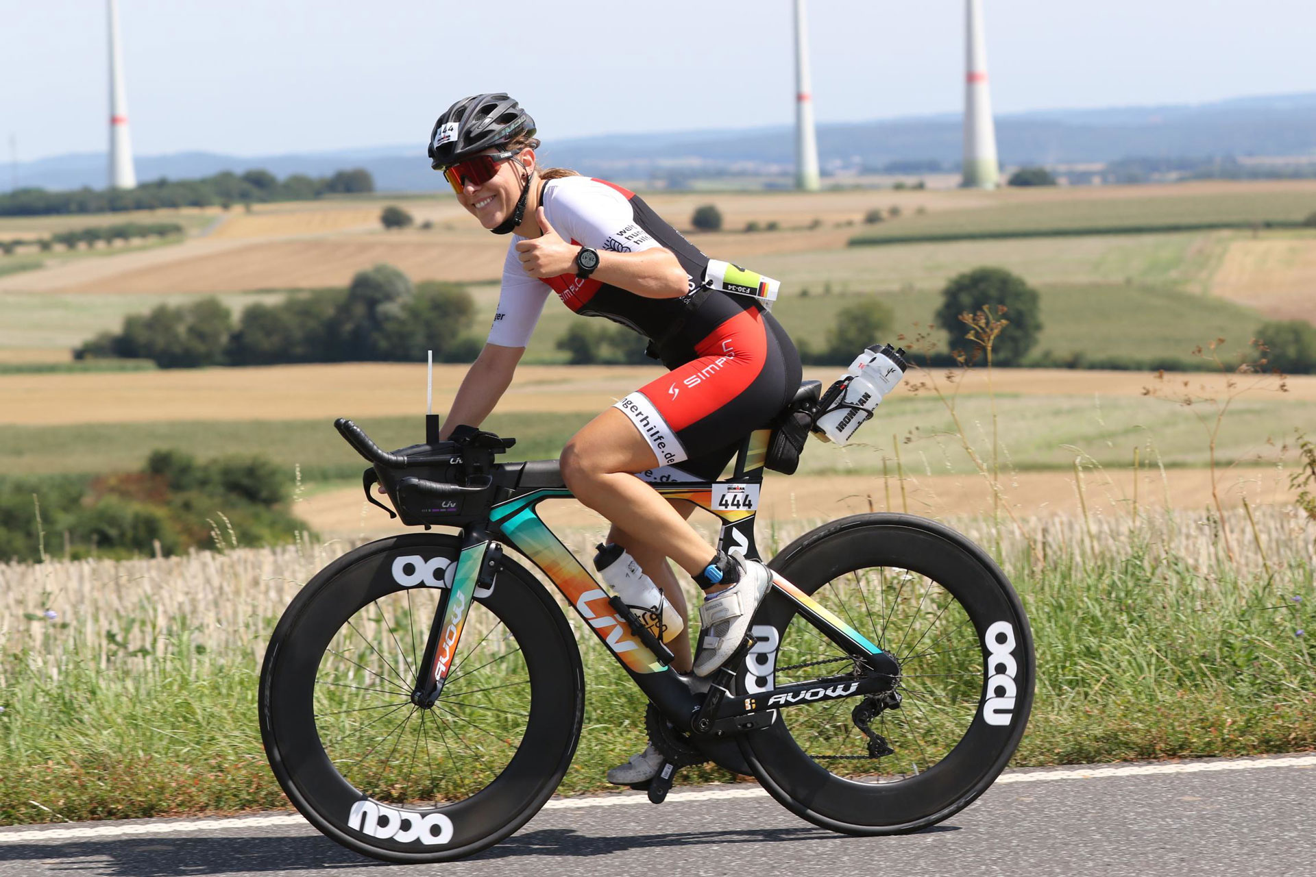 Eine Triathletin auf einem Speed-Fahrrad hält ihren Daumen hoch und lächelt in die Kamera. Sie zeigt ihr Trikot und fährt von einer hügeligen Landschaft.