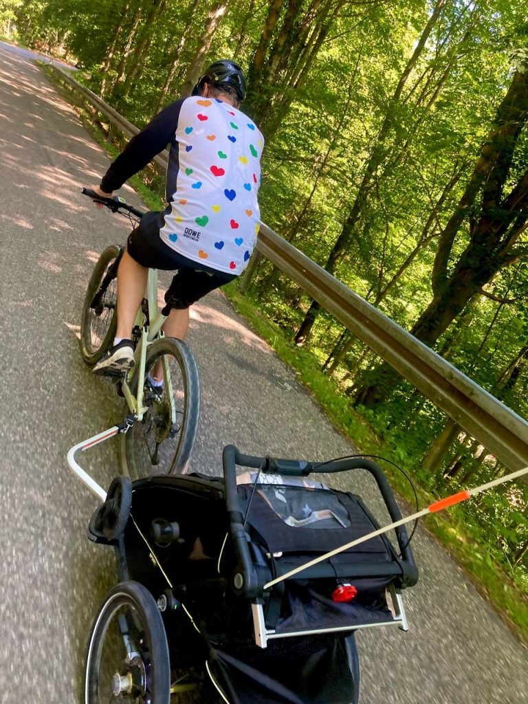 hochzeits-jersey-fahrradanhaenger Ein Bräutigam im Bike-Jersey fährt mit Fahrradanhänger über eine geteerte Straße im Wald. Er ist allerdings nicht als Bräutigam zu erkennen.
