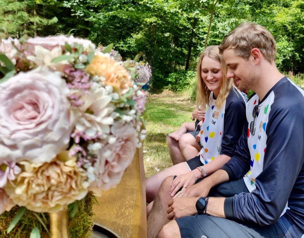 hochzeits-jerseys-bei-trauung Ein Brautpaar Sitzt im Freien auf einer Kirchenbank. Im Vordergrund sind Blumen zu sehen. Das Paar trägt Bike-Jerseys von DOWE Sportswear und umarmt sich.