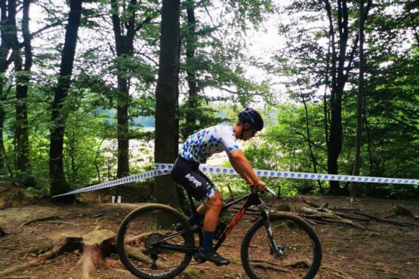 Ein junger Mann im Blau-Weißen Trikot fährt mit dem Mountainbike über einen Waldpfad. Es ist ein Absperrband hinter ihm zu sehen.
