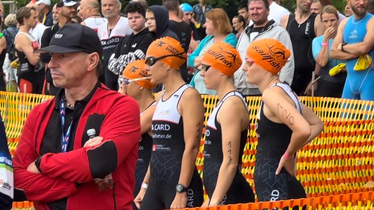 Vier Thiathletinnen in schwarz-weiß-roten Schwimm-Suits, mit oragefarbenen Bademützen und schwarzen Schwimmbrillen. Sie warten vor einem oragefarbenen Absperrband und einer Zuschauermenge. Im Vordergrund steht ein Mann mit rotem Pullover und verschränkten Armen. Er trägt eine schwarze Mütze.