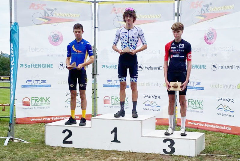 Drei Athleten auf einer Tribüne. Der mittlere, auf dem ersten Platz, ist Leo Hack von den KTM Youngsters. Er trägt ein weißes Trikot von DOWE Sportswear mit dreieck-Muster.