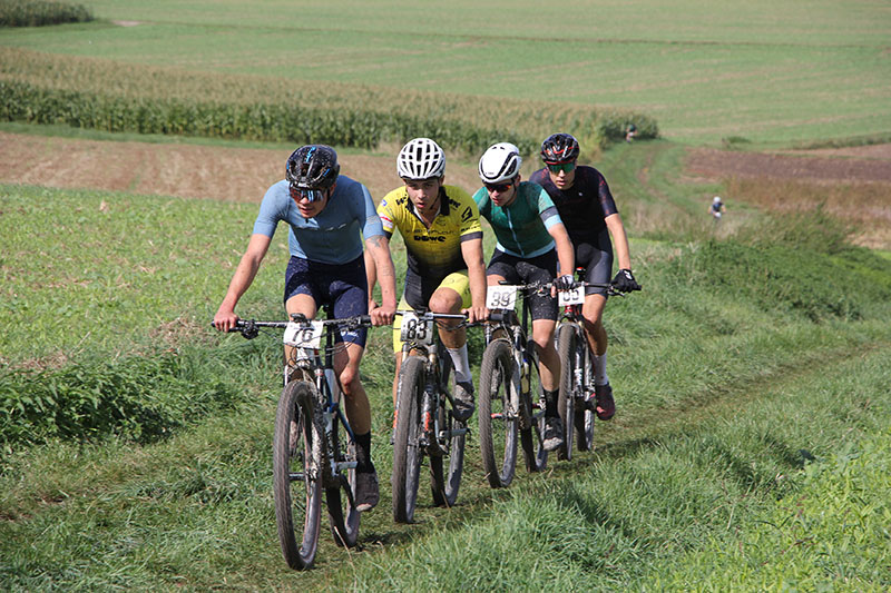 team-dowe-simplon-v10 Vier Mountainbiker fahren hintereinander bei einem Radrennen über eine grasige Landschaft