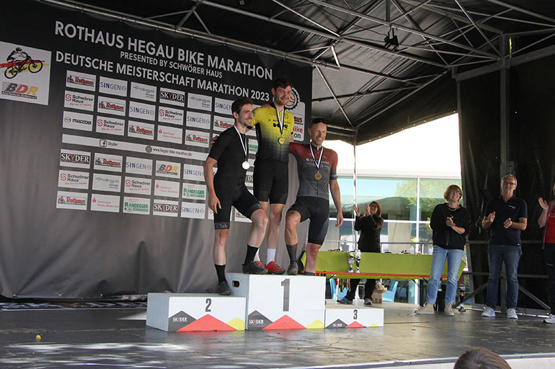 team-dowe-simplon-v14 Drei Radsportler in Trikots auf Tribüne - Auf Platz eins steht Radler im gelben DOWE-Trikot.