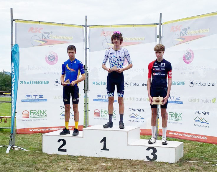 Drei Athleten auf einer Tribüne. Der mittlere, auf dem ersten Platz, ist Leo Hack von den KTM Youngsters. Er trägt ein weißes Trikot von DOWE Sportswear mit dreieck-Muster.